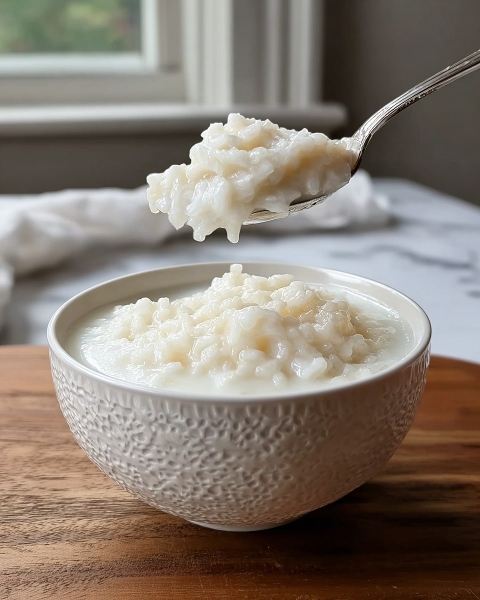 A white textured bowl filled with a creamy, white rice pudding sits on a wooden surface with a white marbled texture in the background. The pudding has a thick, slightly lumpy texture, showing plump grains of rice fully cooked and suspended in a smooth milky liquid. Above the bowl, a silver spoon is lifted, scooping up a generous portion of the rice pudding, highlighting its soft, moist, and shiny appearance. Natural light filters from a window in the background, softly illuminating the dish with a fresh, homey feel. Photo taken with an iphone --ar 4:5 --v 7