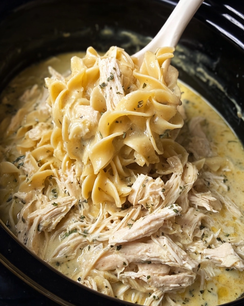 A close-up image of creamy chicken noodle soup inside a black slow cooker. The dish mainly shows soft, thick pasta noodles in a light yellow sauce that looks smooth and glossy, mixed with several pieces of shredded white chicken meat layered evenly throughout. Some small green herb flakes and black pepper specks are scattered around the noodles and chicken, adding fine texture contrast. A white ladle is partially submerged on the right side, holding a serving of the soup above the pot, showcasing the thick and hearty consistency of the dish. photo taken with an iphone --ar 4:5 --v 7