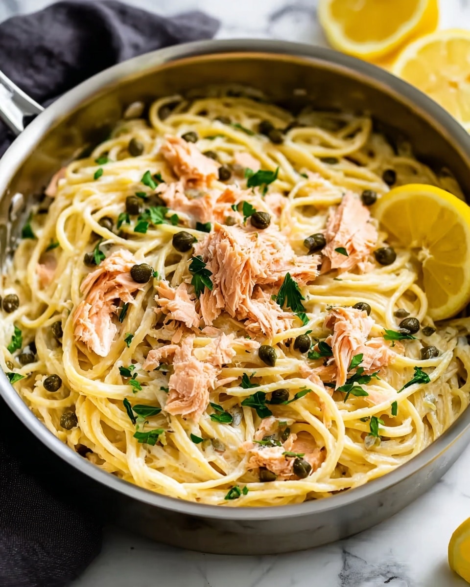 The image shows a white bowl filled with creamy spaghetti pasta topped with several pieces of pink, flaky cooked salmon. Scattered dark green capers and small green parsley leaves are sprinkled on top, adding a fresh color contrast. A bright yellow lemon wedge is placed on the side inside the bowl, resting on the pasta. The bowl is set on a white marbled surface with some lemon wedges nearby. The creamy sauce on the pasta looks smooth and coats the noodles well, giving the dish a rich and inviting appearance. photo taken with an iphone --ar 4:5 --v 7