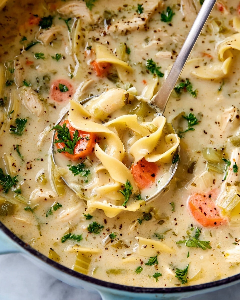A close-up of a white bowl filled with thick creamy chicken noodle soup, showing a mix of wide egg noodles, chunky orange carrot slices, light green celery pieces, and shredded chicken, all in a rich light yellow broth. The soup is topped with finely chopped green herbs, scattered evenly across the surface, with a wooden spoon resting inside the bowl. The bowl is placed on a white marbled surface. Photo taken with an iphone --ar 4:5 --v 7