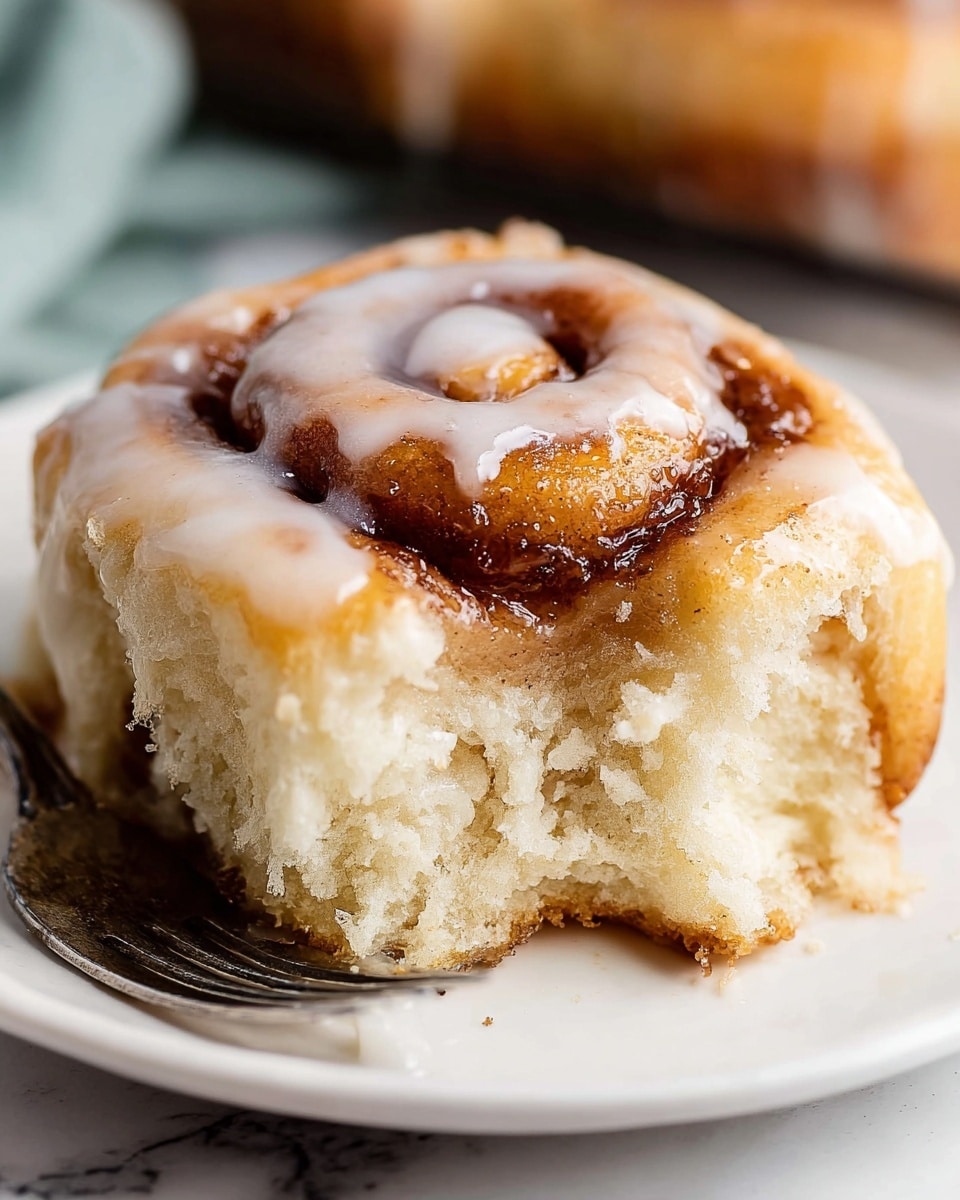 A close-up view of a cinnamon roll resting on a white plate with a small chunk torn off in the front, revealing a soft, fluffy, and light cream-colored inner texture. The cinnamon roll has one main visible spiral layer, with a glossy, golden-brown baked outer part lightly drizzled with white icing that adds a shiny, smooth texture. At the bottom right, a silver fork is partially visible, touching the torn piece. The background shows a white marbled texture. photo taken with an iphone --ar 4:5 --v 7