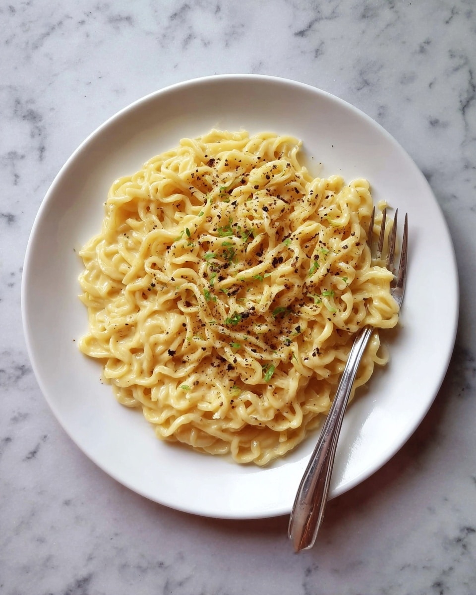 A white plate filled with a single thick layer of creamy yellow noodles mixed evenly, topped with small bits of green herbs and black pepper sprinkled across the center. A silver fork rests along the right edge of the plate. The plate sits on a white marbled texture surface. photo taken with an iphone --ar 4:5 --v 7