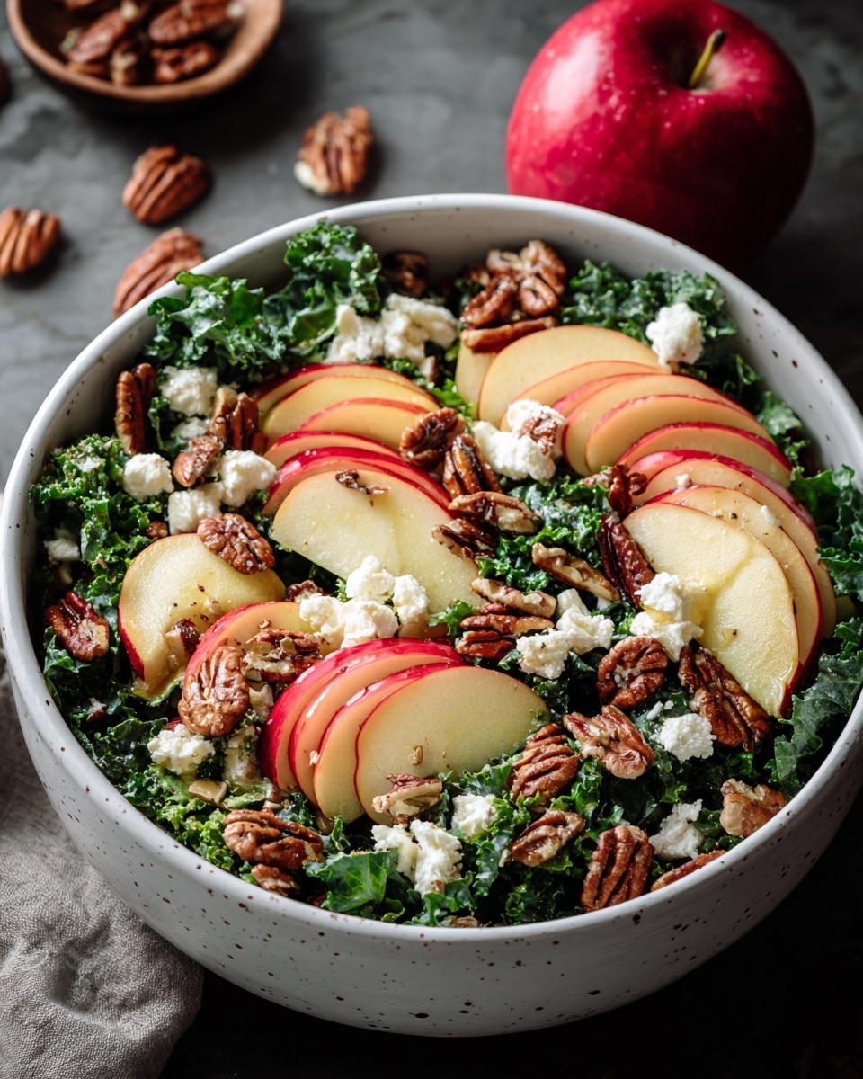 A bowl of fresh salad with three clear layers is shown: the bottom layer is dark green curly kale leaves, the middle layer has thin slices of red and yellow apple with smooth skin, and the top layer is sprinkled with brown, textured pecan nuts and small white chunks of soft cheese. The bowl is white with subtle speckles, sitting on a white marbled surface. In the background, there is a red apple and scattered pecans. Photo taken with an iphone --ar 4:5 --v 7