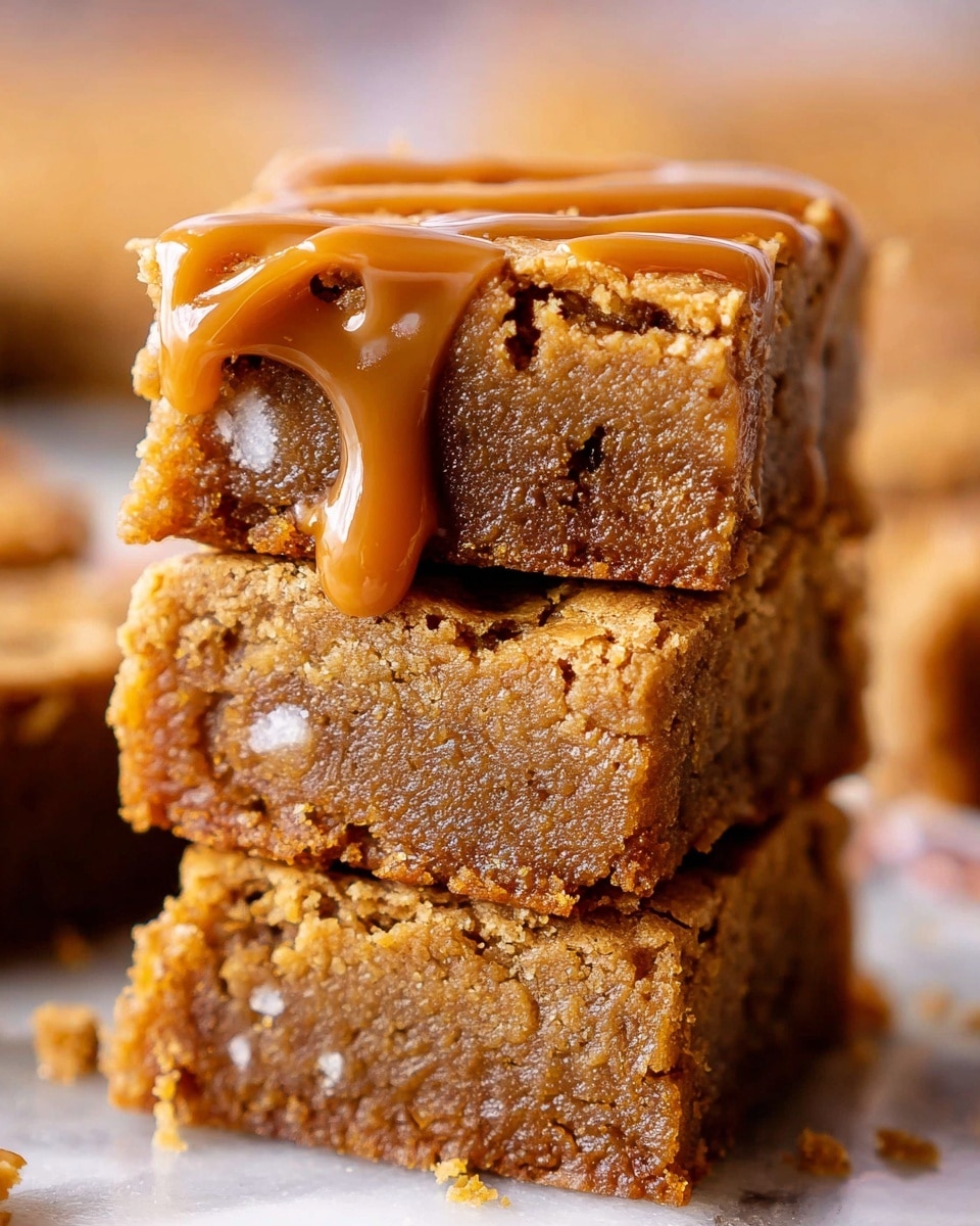 The image shows a close-up of a rectangular cookie held by two woman's hands with neatly painted light nails. The cookie has a golden-brown color with visible chocolate chips embedded throughout. A smooth, thick layer of creamy caramel sauce covers the entire top surface of the cookie, giving it a shiny texture. The background is softly blurred but features a white marbled texture and hints of other cookies. Photo taken with an iphone --ar 4:5 --v 7