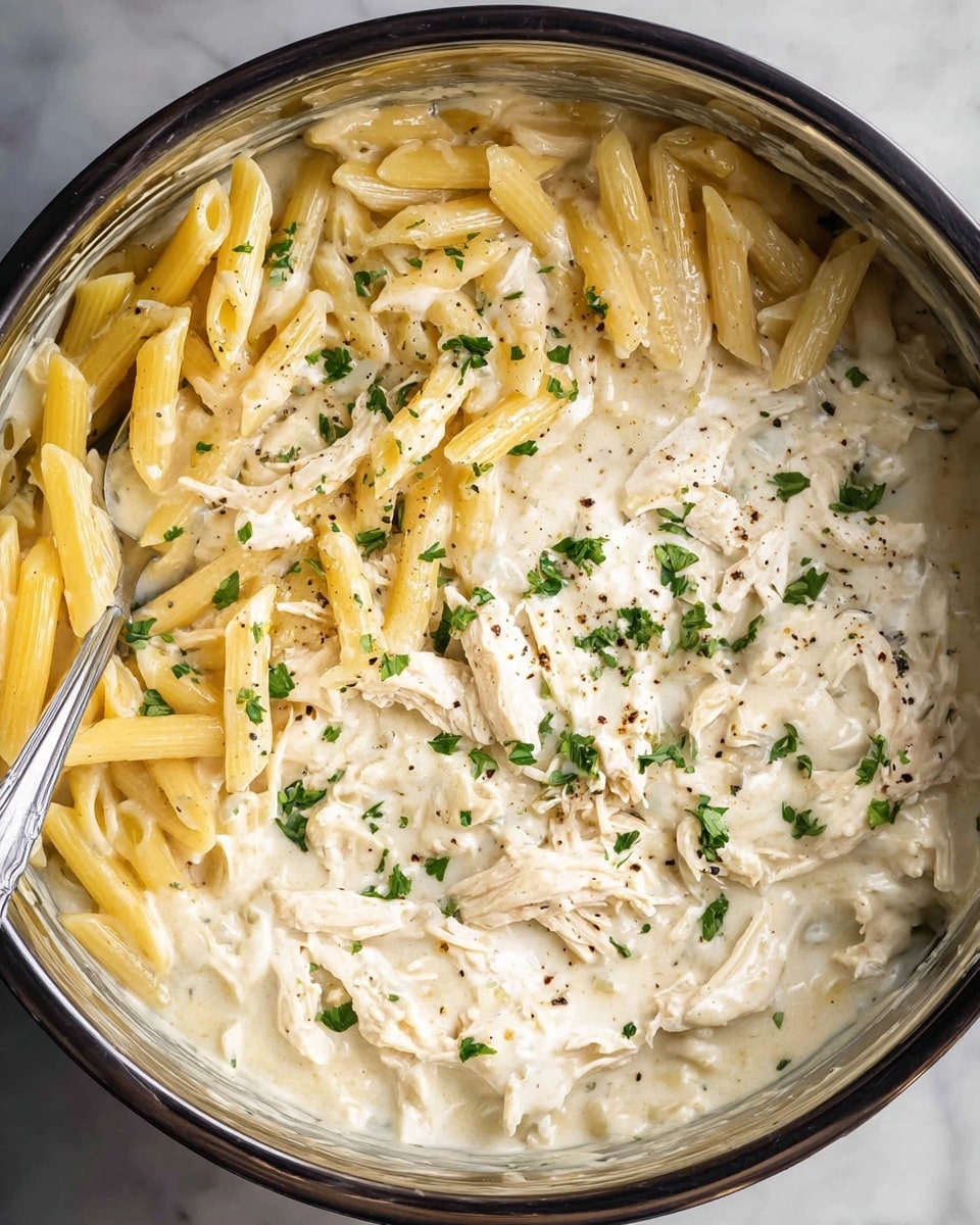 This image shows a close-up view of creamy pasta in a cooking pot. The dish has two main layers: the top layer is creamy white sauce mixed with shredded white chicken pieces and some penne pasta in light yellow color. The sauce is thick and smooth, covering much of the pasta and chicken. Green chopped herbs and small black pepper flakes are scattered evenly on top, adding a touch of color contrast. The metal spoon is partially dipped into the creamy mix on the left side of the pot. The pot is sitting on a white marbled surface. photo taken with an iphone --ar 4:5 --v 7