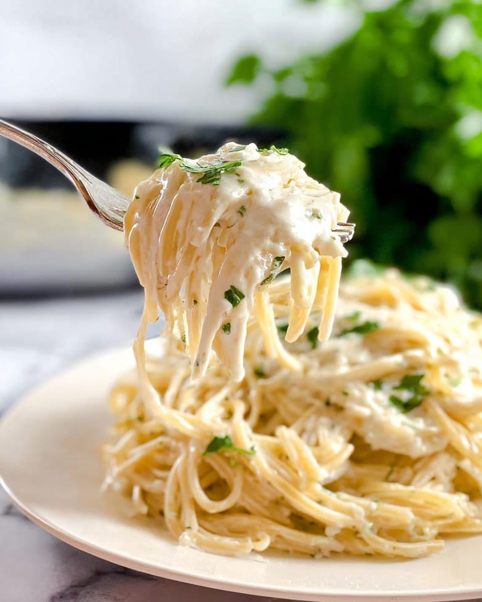A close-up of spaghetti coated in a creamy white sauce with small green herb pieces sprinkled on top, lifted by a fork above a pile of the same spaghetti on a white plate, all set on a white marbled surface with green blurred leaves in the background. Photo taken with an iphone --ar 4:5 --v 7