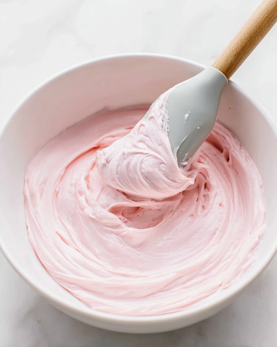 A white bowl holds a smooth, creamy light pink frosting with soft swirls and peaks visible in the texture. A spatula with a wooden handle and a pale gray silicone head is partially dipped into the frosting, lifting a dollop that curls on the edge, showing the thick, airy consistency. The background is a white marbled surface adding a clean, bright look to the image. photo taken with an iphone --ar 4:5 --v 7