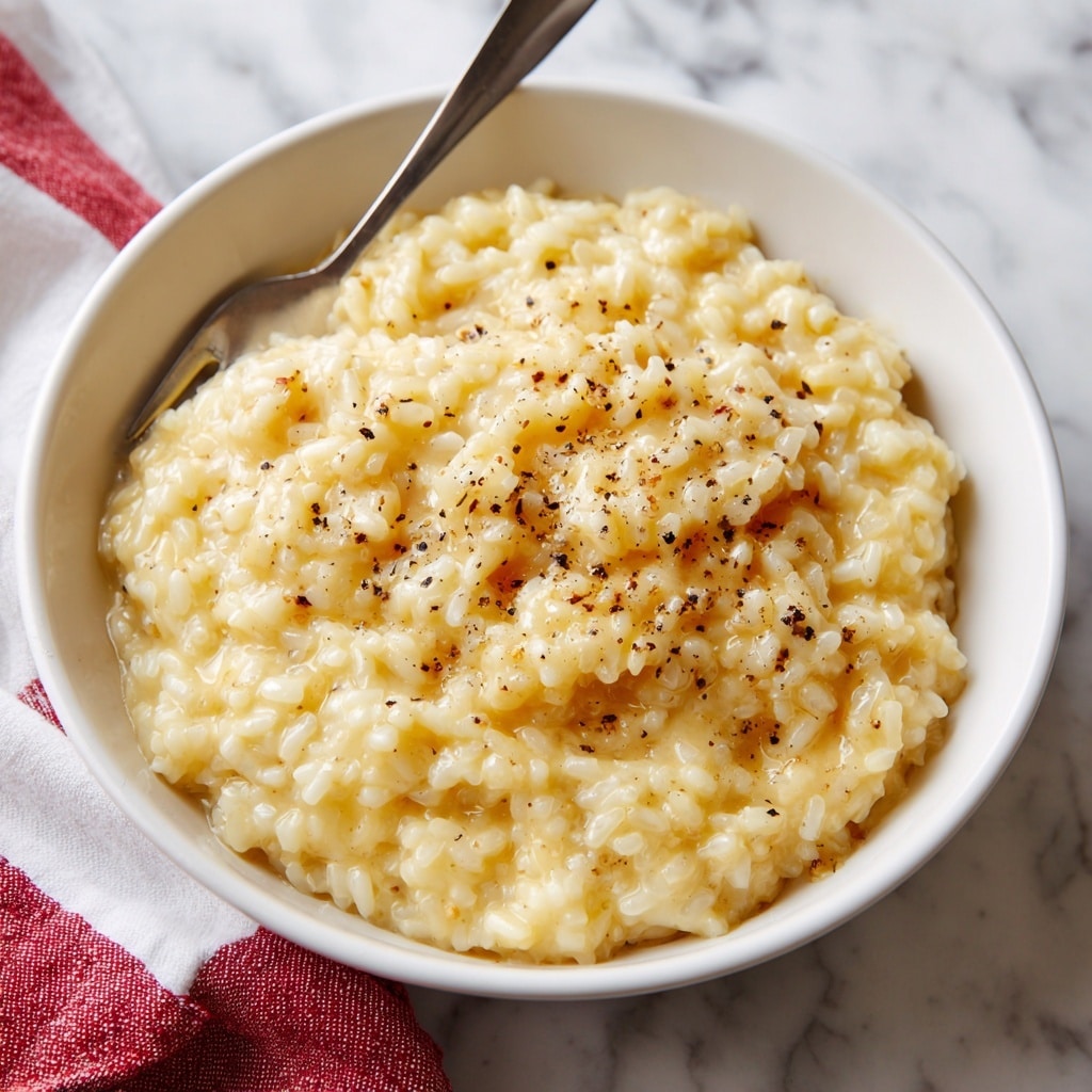 A white bowl filled with creamy, cheesy yellow risotto that has visible grains of soft rice mixed throughout. The risotto has a smooth texture with melted cheese giving it a rich, slightly shiny look. Small black pepper specks are scattered on top, adding contrast. A silver fork is inserted into the risotto on the left side of the bowl. The bowl sits on a white marbled surface with a blurred kitchen towel in the background. Photo taken with an iphone --ar 4:5 --v 7