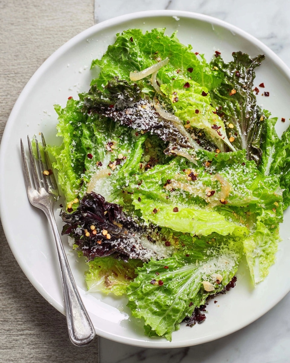 A white plate holds a fresh green salad with two main layers of leafy greens: the bottom layer is made of light green lettuce with soft, smooth leaves, while the top layer has darker green leaves with red edges and a slightly rough texture. Both layers are sprinkled with white grated cheese and small red chili flakes, adding spots of color across the greens. A silver fork rests on the left side of the plate, slightly touching the salad. The plate sits on a white marbled surface with another plate of similar salad blurred in the background. photo taken with an iphone --ar 4:5 --v 7