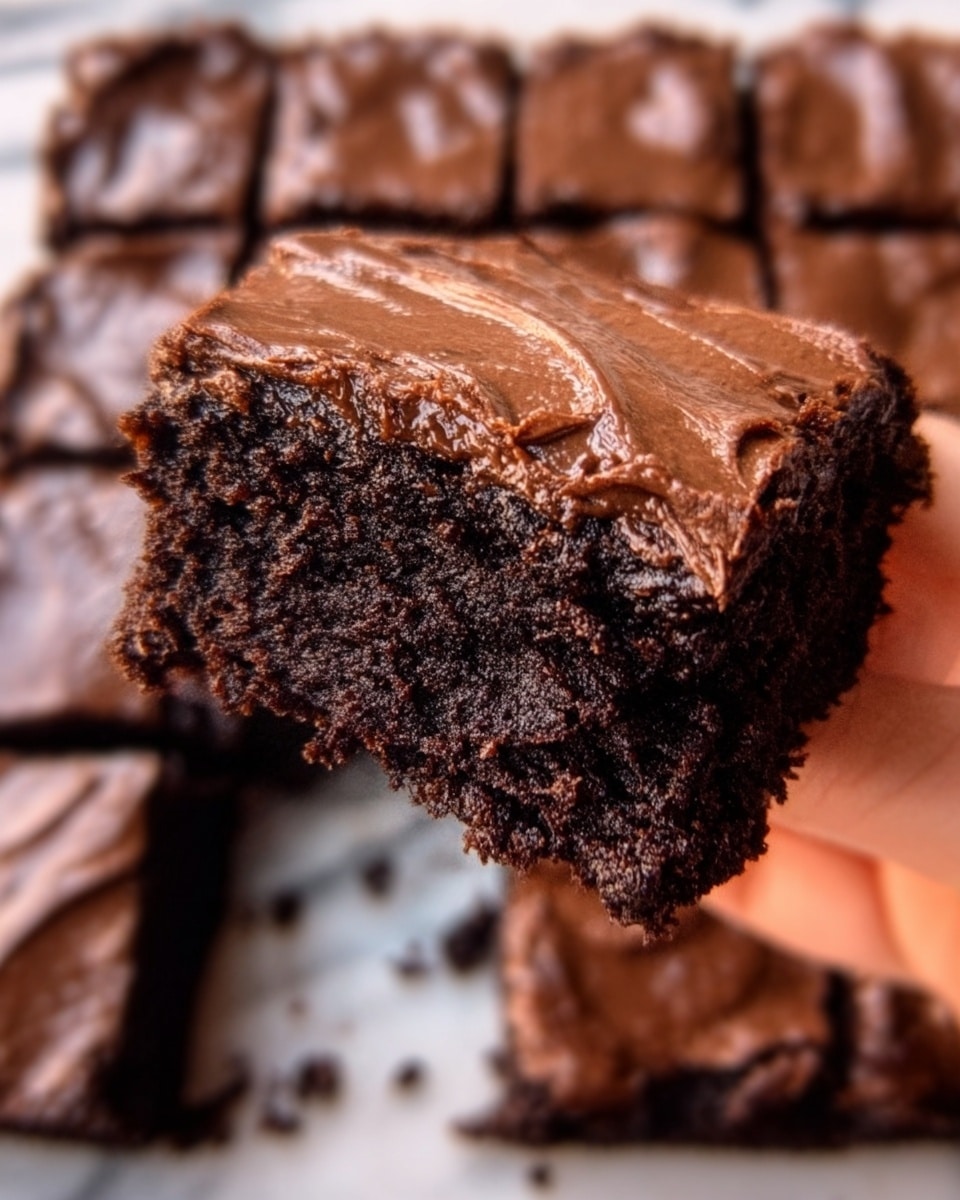 A close-up image of a thick, square piece of chocolate brownie being held by a woman's hand. The brownie has a dark, rich, and slightly crumbly texture with a smooth, shiny chocolate layer spread thickly on top. The background shows more brownies cut into square pieces, placed on a white marbled surface, creating a uniform grid pattern. The lighting highlights the moist and dense texture of the brownie and the glossy finish of the chocolate topping. Photo taken with an iphone --ar 4:5 --v 7