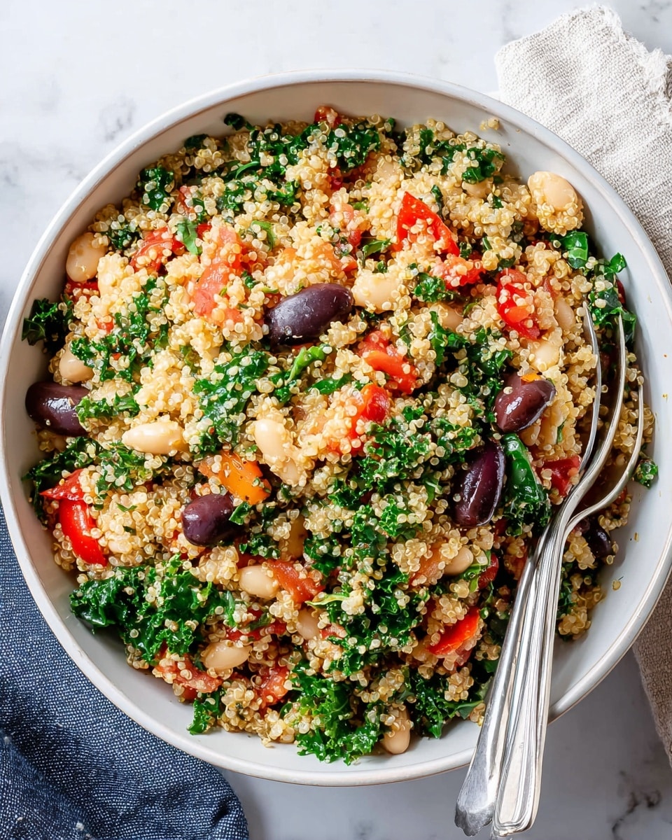 A dark blue bowl filled with a mixed quinoa salad, showing multiple layers including light yellow quinoa grains, dark purple olives, white beans, bright red diced tomatoes, and dark green chopped kale all evenly mixed together. Two silver spoons rest inside the bowl on the left side. The bowl is placed on a white marbled surface next to a beige linen cloth on the right side. photo taken with an iphone --ar 4:5 --v 7