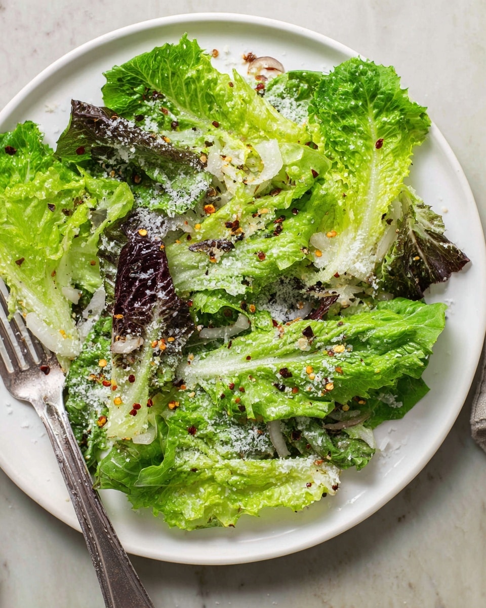 A white plate holds a fresh salad with layers of green and dark purple lettuce leaves as the base, topped with a light sprinkle of grated cheese and scattered red chili flakes. The lettuce looks crisp and moist with different textures, some ruffled, some smooth. Thin slices of shallots are mixed in lightly visible under the leaves. A silver fork rests on the left edge of the plate, placed on a white marbled surface. photo taken with an iphone --ar 4:5 --v 7