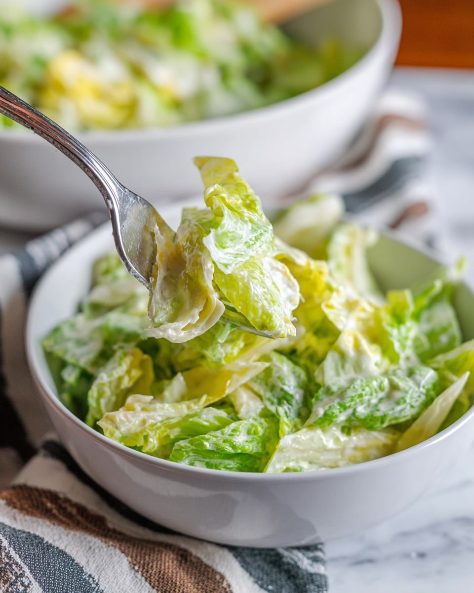 A white oval bowl filled with a base layer of bright green chopped iceberg lettuce, fresh and crisp with visible veins and texture. On top, there is a thick layer of creamy, white ranch dressing with visible small chunks and specks of seasoning, unevenly spread across the lettuce in wide dollops. The bowl is placed on a white marbled surface. photo taken with an iphone --ar 4:5 --v 7