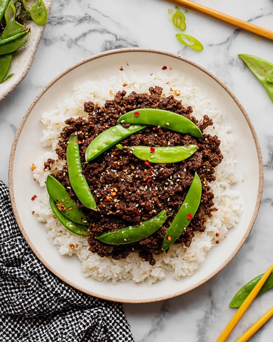 A white round plate holds a dish with two main layers: the bottom layer is fluffy white rice spread evenly around the plate's edge, and the top layer is dark brown ground meat mixed with bright green sugar snap peas scattered on top. The meat is sprinkled with small white sesame seeds and some red chili flakes for color. The plate sits on a white marbled surface with a small white dish containing red chili flakes partially visible to the side. photo taken with an iphone --ar 4:5 --v 7