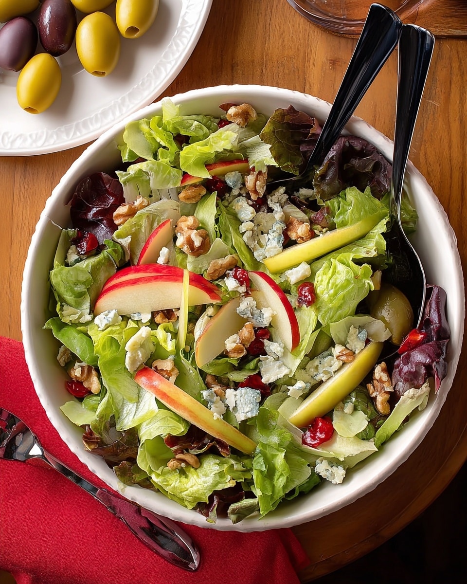 A large white bowl filled with a fresh salad featuring a mix of green and purple leafy lettuce as the base layer, topped with slices of red apple and green pear scattered evenly on top. Small chunks of blue cheese and chopped walnuts add texture, with pieces of red bell pepper scattered throughout. Two black salad utensils rest inside the bowl. The bowl sits on a wooden table with a red napkin to the left and a red container with a light-colored liquid in the top right corner, all set against a white marbled texture background. Photo taken with an iphone --ar 4:5 --v 7