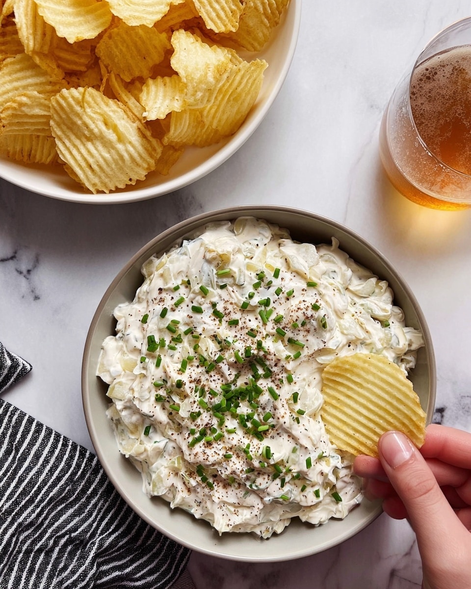 A large bowl of creamy white dip with small pieces of cooked onion mixed in, topped with finely chopped green chives and a sprinkle of black pepper on top, with a ridged potato chip dipped into the side of the bowl. Nearby, a white bowl full of more ridged potato chips sits above it on a white marbled surface, along with a glass of light amber drink to the right on a striped black and white cloth. A woman’s hand is holding one chip in the dip, photo taken with an iphone --ar 4:5 --v 7