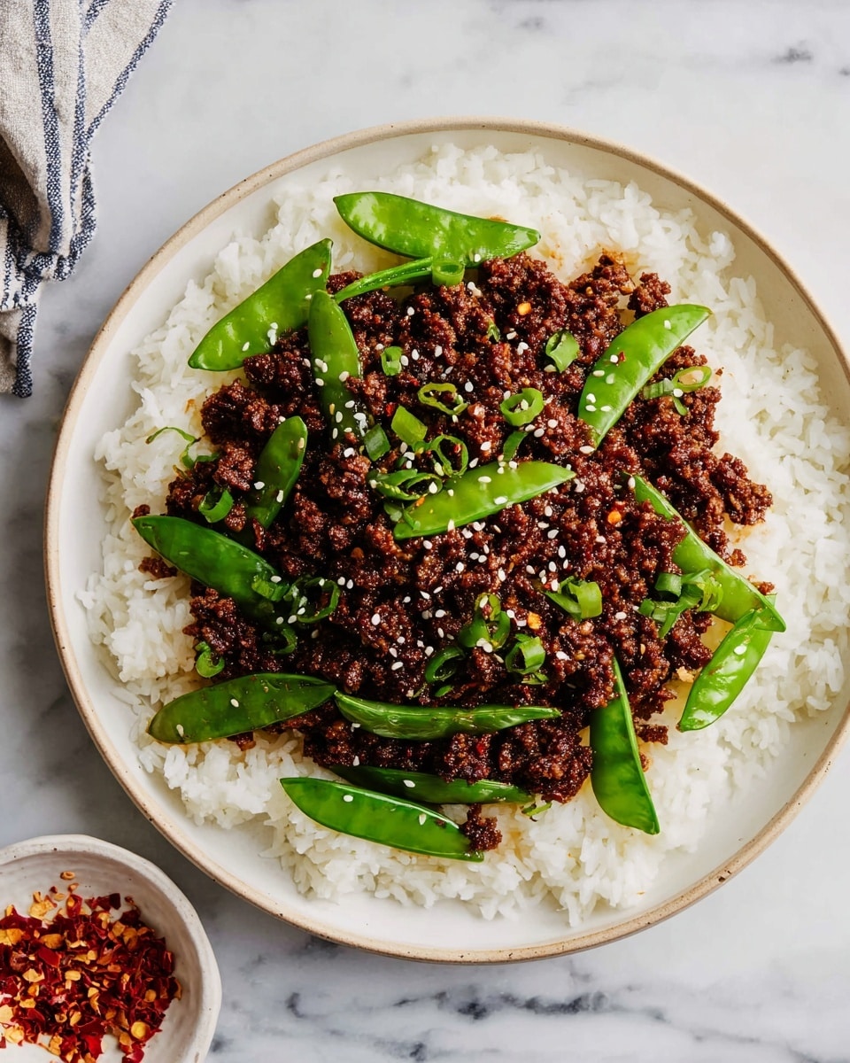 The dish shows a white plate filled with a base layer of white rice with a soft, fluffy texture spread evenly. On top, there is a thick layer of dark brown, finely ground beef mixed with small bits of seasoning. Nestled in the beef layer are bright green sugar snap peas, whole and fresh, adding a smooth and glossy texture. Some white sesame seeds and red chili flakes are sprinkled lightly over the beef and peas for a touch of contrast. The plate sits on a white marbled surface, next to a black and white checkered cloth and yellow chopsticks. Photo taken with an iphone --ar 4:5 --v 7