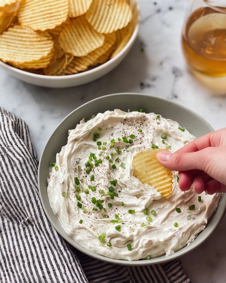 A close-up image of a creamy white dip with a thick, smooth texture, swirled in a simple grey bowl. The dip is topped with small green chive pieces and a sprinkling of coarse black pepper, adding texture and color contrast. A woman's hand is dipping a ridged, pale yellow potato chip into the dip, slightly pressing into it. Behind the bowl of dip is a white bowl filled with more ridged potato chips, and to the right is a glass of light amber liquid. The setting is on a white marbled surface with a black and white striped cloth underneath the bowl of dip. photo taken with an iphone --ar 4:5 --v 7