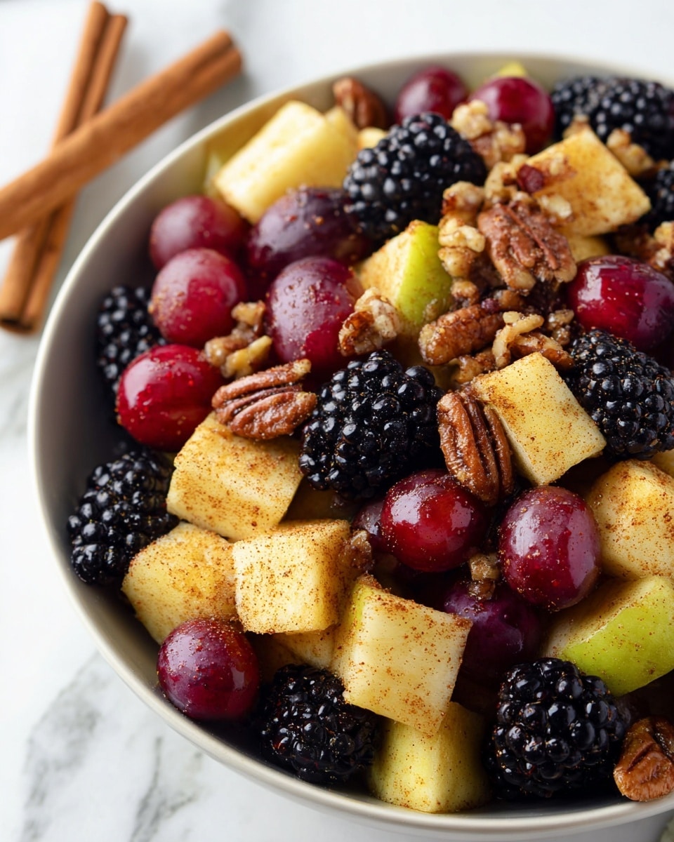 A close-up view of a white bowl filled with a fresh fruit salad consisting of three layers. The bottom layer is made up of dark blackberries, the middle layer includes glossy red grapes, and the top layer has yellowish cubed apples sprinkled with a light dusting of cinnamon and mixed with scattered pieces of brown walnuts and pecans. The bowl is placed on a white marbled surface with two cinnamon sticks nearby and a silver whisk partially visible in the background with a red patterned cloth draped beside it. photo taken with an iphone --ar 4:5 --v 7