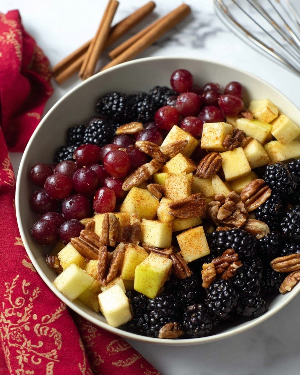 A close-up view of a bowl filled with a colorful fruit salad, showing three main layers: the bottom layer is shiny black blackberries, the middle layer has large red and dark purple grapes, and the top layer features yellowish apple chunks dusted with cinnamon and scattered brown pecans. The fruits look fresh and slightly glossy, with the apple pieces having a grainy spice coating. The bowl itself is white, placed on a white marbled surface, and two cinnamon sticks lie next to it. Photo taken with an iphone --ar 4:5 --v 7