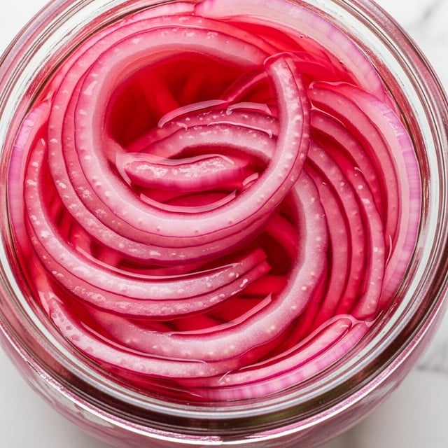 A close-up image of a glass jar filled with thinly sliced pickled red onions soaking in a vivid pinkish-red liquid. The onion slices are translucent with a mix of soft pink and deeper red hues, showing their smooth, slightly shiny texture as they curve and stack closely inside the jar. The jar rim is visible, and the background shows a white marbled texture. photo taken with an iphone --ar 4:5 --v 7