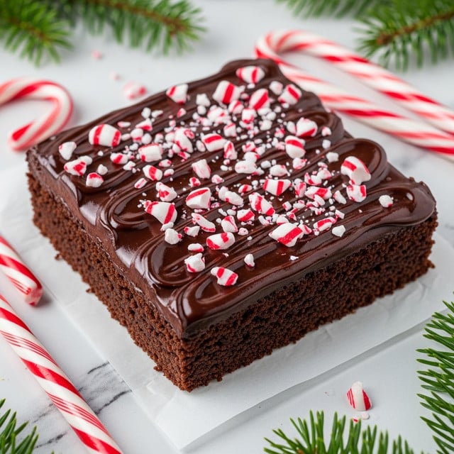 A square-shaped chocolate brownie forms the base layer, dark brown with a slightly rough texture on the sides. The top is covered in a thick, glossy dark chocolate ganache layer that spreads unevenly across the surface with shiny, wavy strokes. Scattered generously over the ganache are broken pieces of red and white striped peppermint candy canes, adding a bright, festive contrast. The brownie sits on a piece of parchment paper placed on a white marbled surface. Around the brownie are whole candy canes and sprigs of green pine, adding to the holiday vibe. Photo taken with an iphone --ar 4:5 --v 7