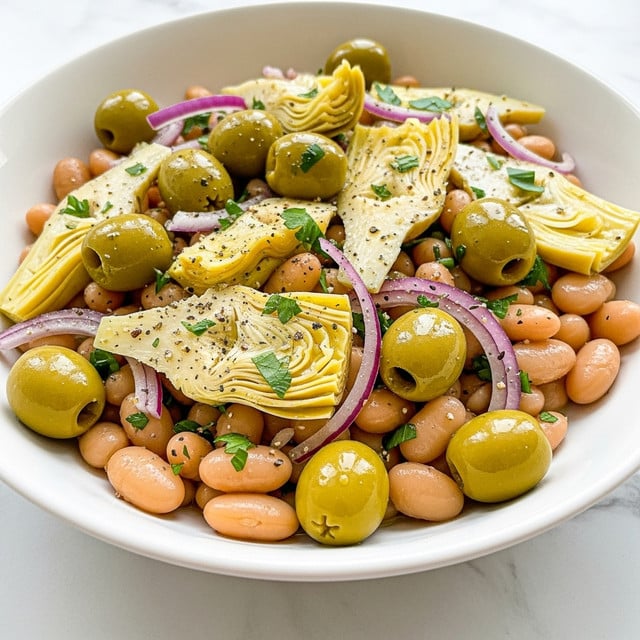 A close-up view of a white bowl filled with a colorful bean salad. The salad has three main layers: at the bottom and mixed throughout are light beige beans with smooth, round shapes; scattered among them are bright green whole olives, glossy and firm; on top and throughout are slices of light yellow artichoke hearts with a soft texture, thin slices of purple-red onion, and chopped dark green herbs sprinkled all over. The salad is lightly coated in a glistening olive oil dressing, with small specks of black pepper and other seasonings visible. The bowl sits on a white marbled surface. photo taken with an iphone --ar 4:5 --v 7