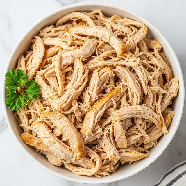 A white bowl filled with shredded cooked chicken, showing many thin layers of light beige and golden brown meat with a slightly dry texture. The chicken pieces are uneven in size, piled loosely, and a small sprig of green parsley is placed on the left side of the bowl for garnish. The bowl is set on a white marbled surface with a hint of a striped cloth at the bottom right edge of the image. The photo taken with an iphone --ar 4:5 --v 7