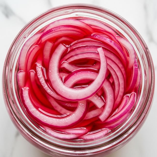 A close-up view of a glass jar filled with thinly sliced pickled red onions submerged in a bright pink liquid. The onion slices are shiny and wet, showing a mix of translucent pale pink and deeper red colors with a slightly curved and soft texture. The jar is open at the top, revealing the packed layers of onions inside. The background is a white marbled texture. photo taken with an iphone --ar 4:5 --v 7