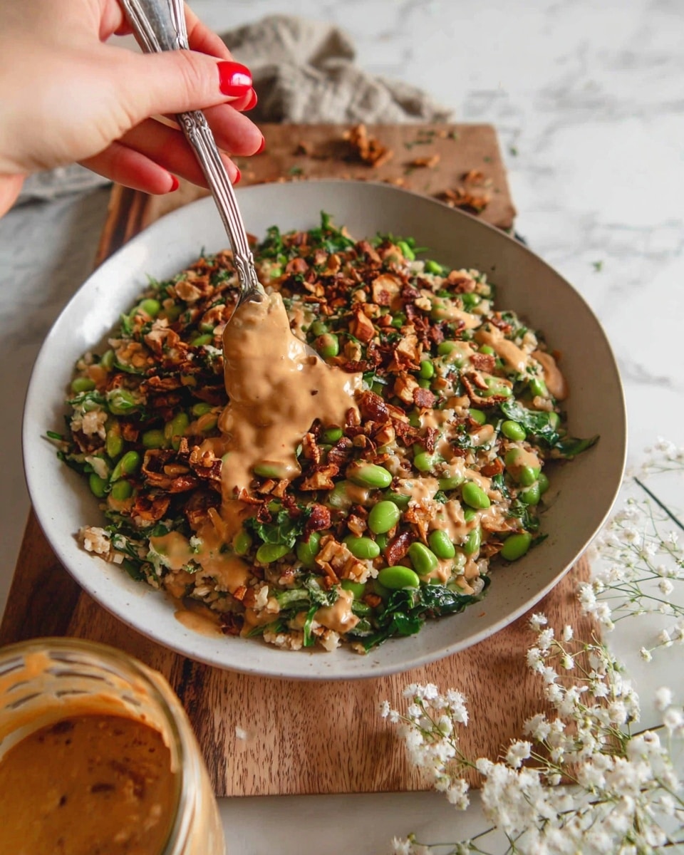 The dish is shown in a shallow white bowl filled with a mixture of chopped ingredients layered together: beige cooked grains form the base, mixed with green edamame beans and wilted green leafy vegetables scattered throughout; on top, there are pieces of browned, crispy nuts, and the whole dish is drizzled with a thick, creamy light brown sauce. A woman's hand with red nail polish holds a silver fork right above the bowl, partially mixing the ingredients. Beside the bowl on a wooden board, there is a glass jar filled with the same sauce with a spoon coated in sauce resting on top. In the background, delicate white flowers add a soft touch. The setting surface has a white marbled texture. photo taken with an iphone --ar 4:5 --v 7
