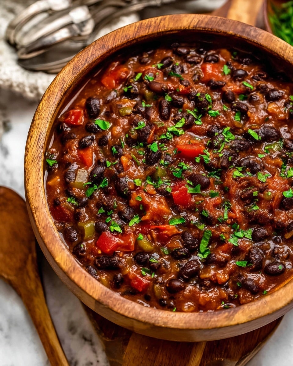 A bowl filled with a hearty black bean stew, showing one thick layer composed of black beans, small red bell pepper pieces, diced tomatoes, and bits of onion mixed in a rich brown sauce. The stew is topped with scattered fresh chopped green cilantro leaves. A silver spoon rests inside the bowl, partially submerged in the stew. The bowl is white with a thin brown rim, placed on a white marbled surface. Nearby, there is a small white bowl with lime wedges and another small white bowl filled with chopped cilantro on a beige cloth. Photo taken with an iphone --ar 4:5 --v 7