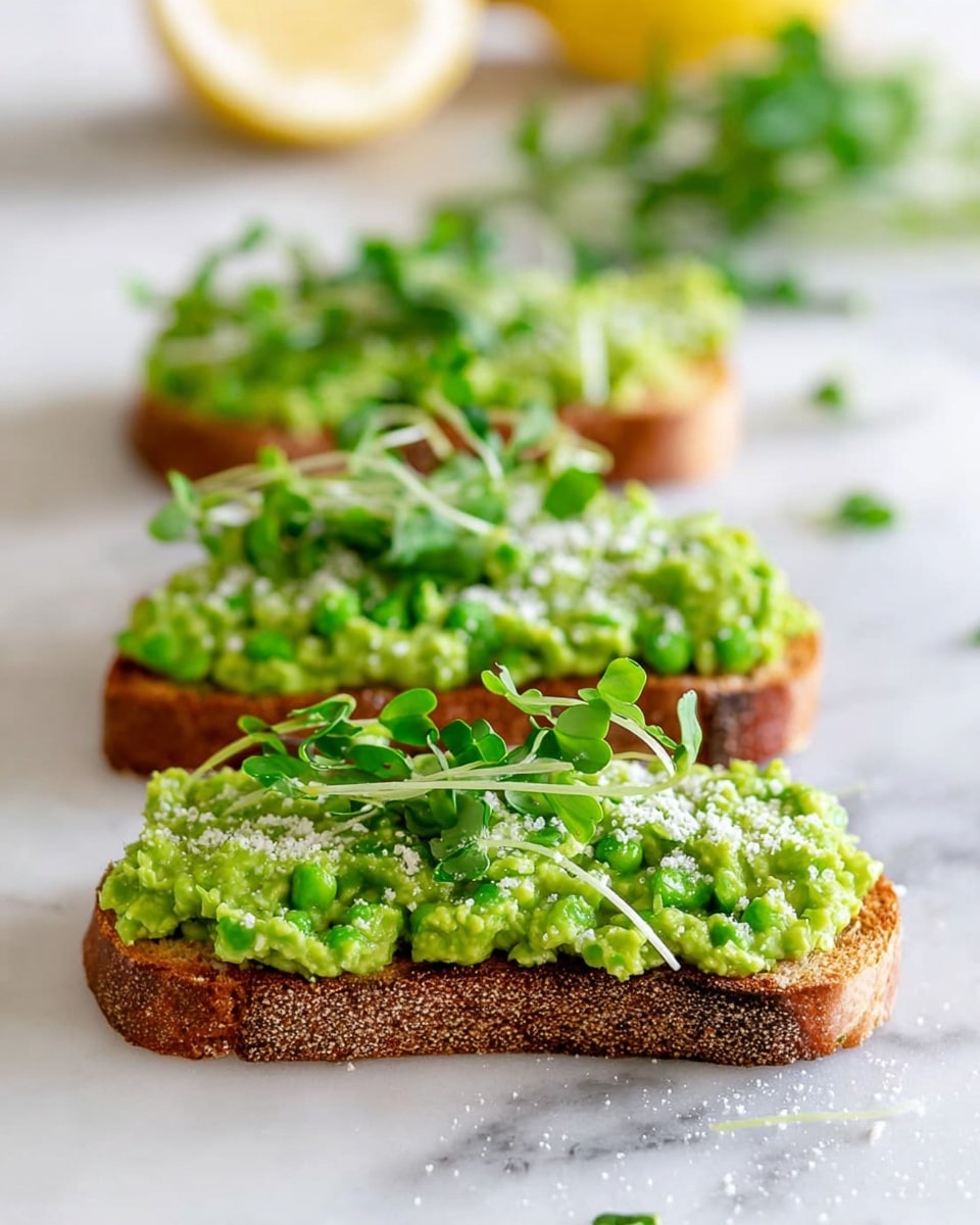 Three slices of toasted brown bread are placed in a row on a white rectangular plate, each slice topped with a thick, chunky layer of bright green mashed avocado mixed with small bits of seasoning. On top of each avocado layer, there is a light sprinkling of white crumbled cheese, adding a touch of texture, and fresh green leafy pea shoots arranged delicately across the surface. In the blurred background, a lemon wedge sits on the white marbled surface, hinting at added freshness. The image is sharp in the front and softly blurred as it goes back, capturing the texture of the toast crust and the creamy avocado clearly. photo taken with an iphone --ar 4:5 --v 7