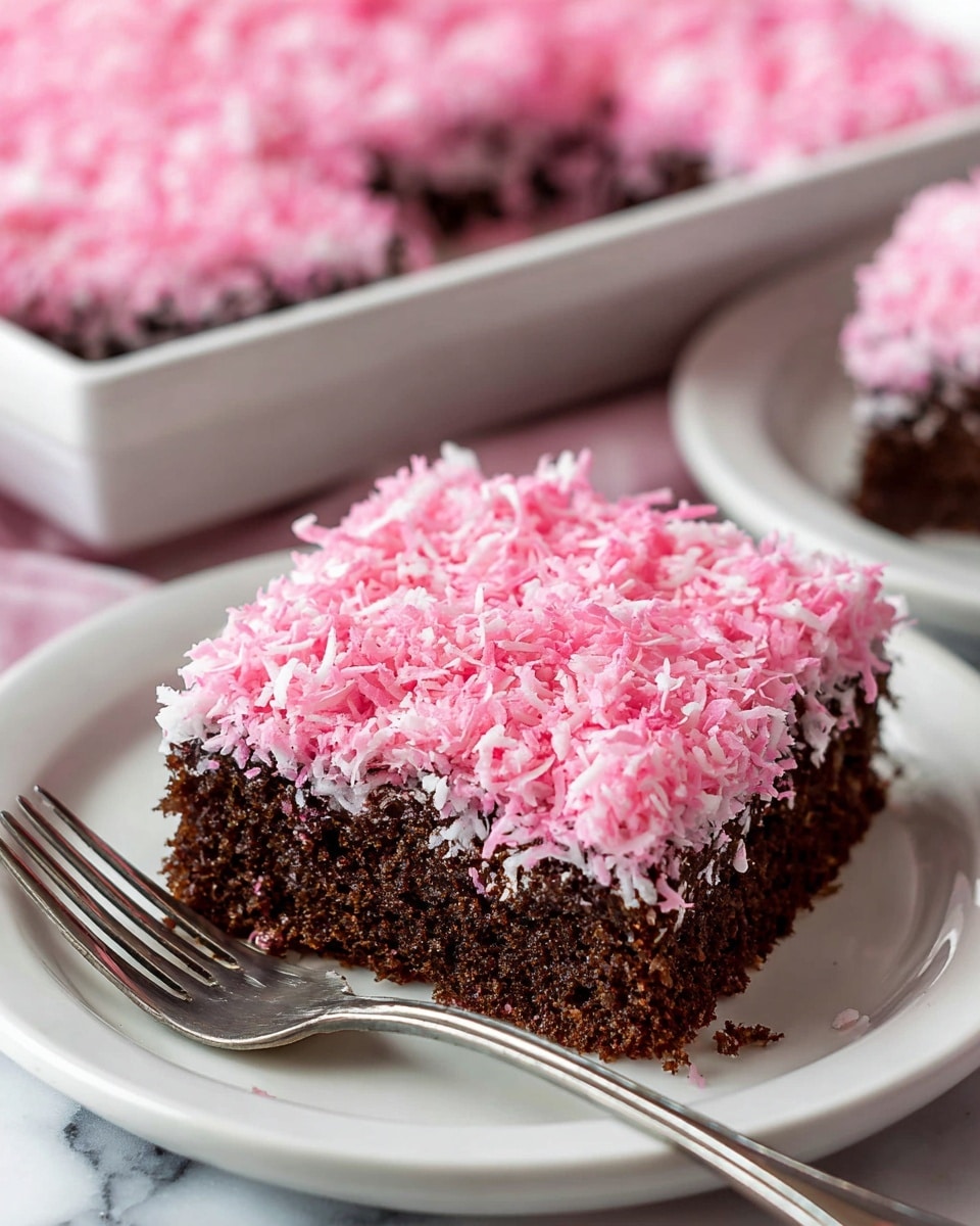 A close-up image shows a square piece of cake on a white plate. The cake has two layers: the bottom layer is dark brown and looks moist with a dense texture, while the top layer is covered with bright pink shredded coconut, giving it a rough and fluffy appearance. The pink topping fully covers the top and sides of the cake slice. A silver fork lies on the plate next to the cake. In the background, a white tray with more of the same cake is slightly blurred, sitting on a white marbled surface. Photo taken with an iphone --ar 4:5 --v 7