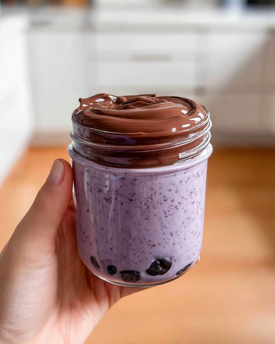 A clear glass jar filled almost to the top with a thick, creamy light purple mixture that has small dark purple specks evenly spread throughout. At the bottom of the jar, there are whole dark purple blueberries pressed against the glass. A smooth, glossy stream of light brown sauce is shown pouring from above into the jar, creating a glossy layer on top of the purple mixture. The jar rests on a surface with a white marbled texture and the background is softly blurred, featuring a person wearing a textured dark blue shirt. photo taken with an iphone --ar 4:5 --v 7