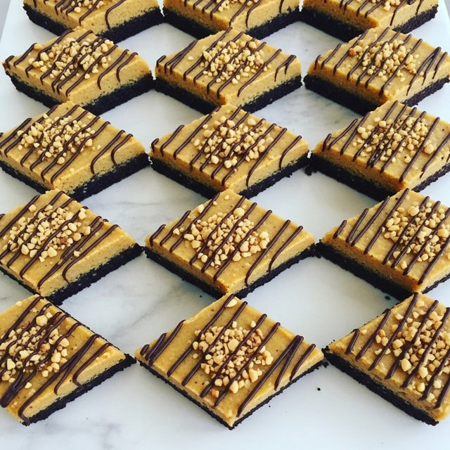 A close-up image of a baking tray filled with 20 diamond-shaped dessert bars, each cut and evenly spaced, resting on a white marbled texture. The bars have a golden-brown top layer with a smooth, slightly creamy texture, sprinkled with chopped nuts in light tan and golden colors. Thin diagonal drizzles of dark chocolate cover the whole tray, creating fine lines that stretch across the dessert bars from edge to edge. The base of the bars looks darker and firm, suggesting a baked crust or brownie-like layer underneath the topping. photo taken with an iphone --ar 4:5 --v 7