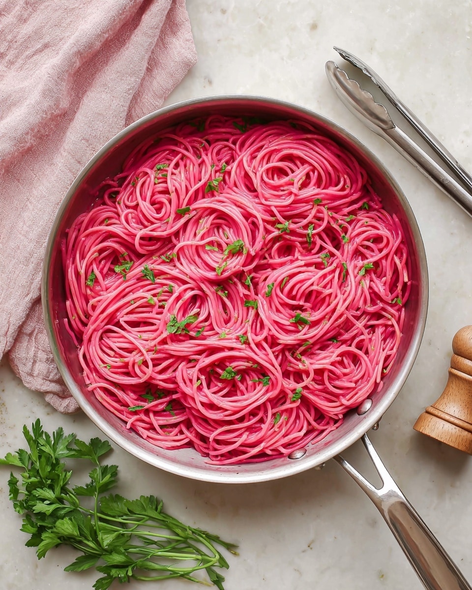A shiny pan filled with bright pink spaghetti, each strand coated evenly in a creamy sauce, is topped with small green parsley pieces scattered across the noodles. The pan is placed on a white marbled surface with a light pink cloth partly visible on the left side, a pair of silver tongs on the right, and a wooden salt or pepper grinder nearby. Some fresh green parsley leaves rest near the pan. The scene has a clean, fresh look. photo taken with an iphone --ar 4:5 --v 7