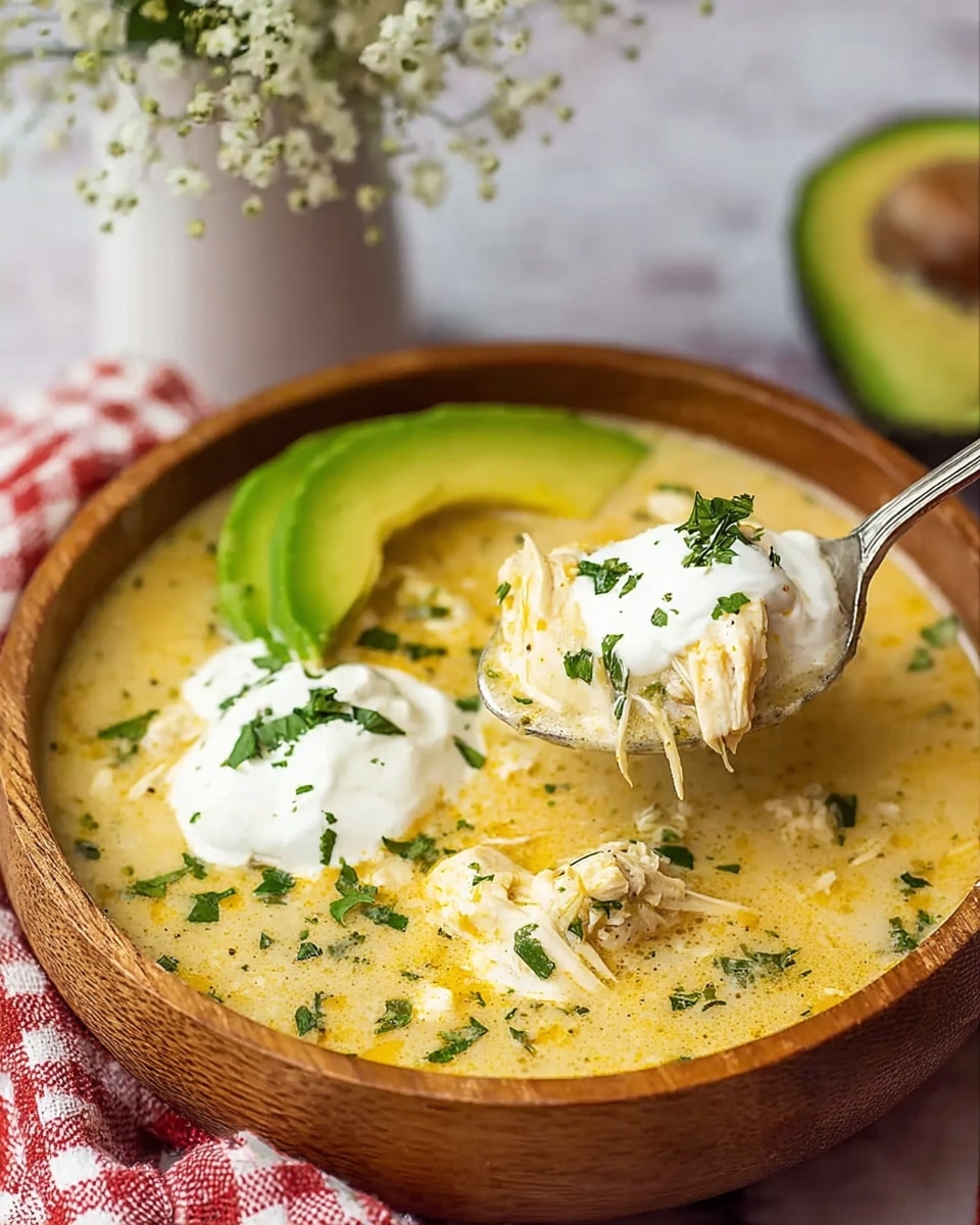 A wooden bowl filled with creamy chicken soup that has a light yellow color with green herb specks throughout, topped with a dollop of white sour cream and sprinkled green herbs. On one side of the bowl, there is a bright green slice of avocado resting on the soup. A silver spoon holds a close-up scoop of the soup showing shredded white chicken meat, some melted yellow cheese, sour cream, and bits of green herbs. The bowl is placed on a white marbled surface near a red and white checkered cloth and a white vase with small white flowers in the background. photo taken with an iphone --ar 4:5 --v 7