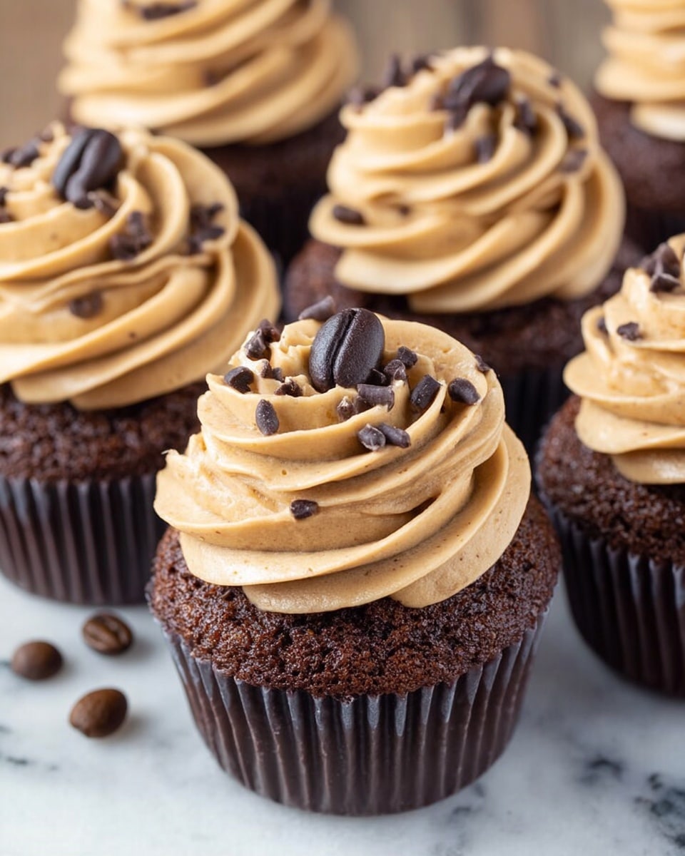 The image shows several chocolate cupcakes arranged close together, each with one layer of moist, dark brown cake at the bottom, topped with a thick swirl of light brown coffee-flavored frosting, which has a smooth and creamy texture. The frosting is decorated with small dark chocolate shavings and a single dark brown coffee bean placed on top, adding detail and contrast. The cupcakes are inside dark brown paper liners, and a few coffee beans lie next to them on a white marbled surface. The photo taken with an iphone --ar 4:5 --v 7