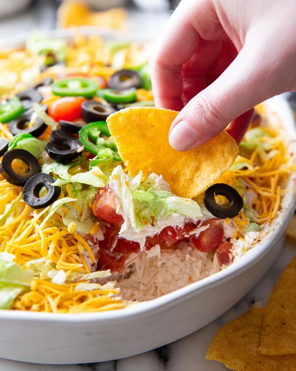 A white dish sits on a wooden board with fresh layered taco salad inside. The bottom layer is green lettuce, followed by bright red diced tomatoes and green sliced jalapeños. On top is a generous layer of shredded yellow and white cheese mixed with black olive slices. Surrounding the dish are small clear bowls with more diced tomatoes, olives, and cheese, plus a glass bowl full of yellow tortilla chips. Some chips are on a white plate nearby, all on a white marbled surface. photo taken with an iphone --ar 4:5 --v 7