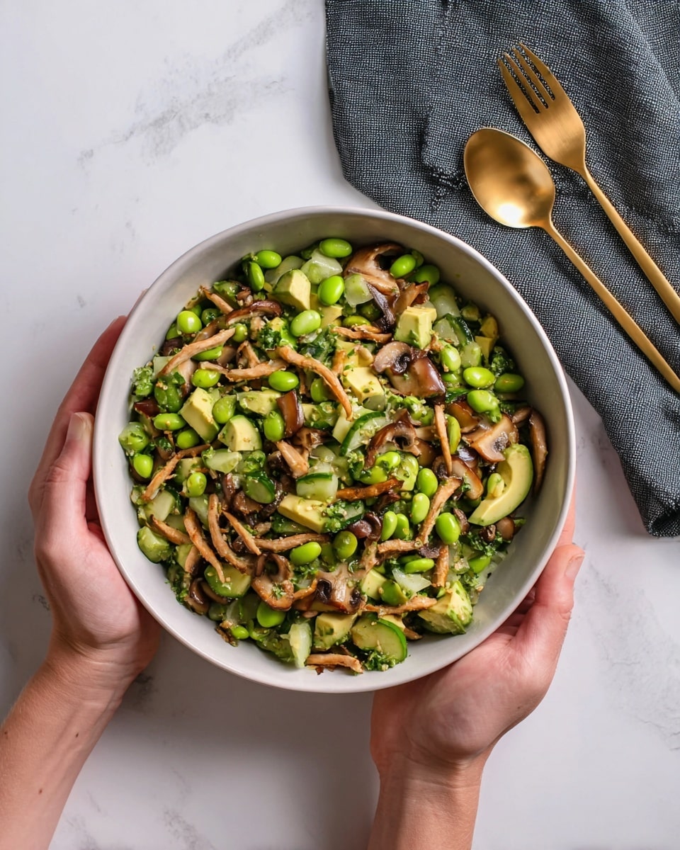 A white bowl filled with a mixed salad held by a woman's hands from the sides. The salad has layers of bright green edamame beans, diced cucumber pieces, creamy light green avocado chunks, and thin, long cooked mushrooms that are golden brown with darker tips. Small green onion slices are spread throughout. The bowl is on a white marbled surface, with a dark grey cloth napkin to the upper right, next to a gold fork and spoon. photo taken with an iphone --ar 4:5 --v 7