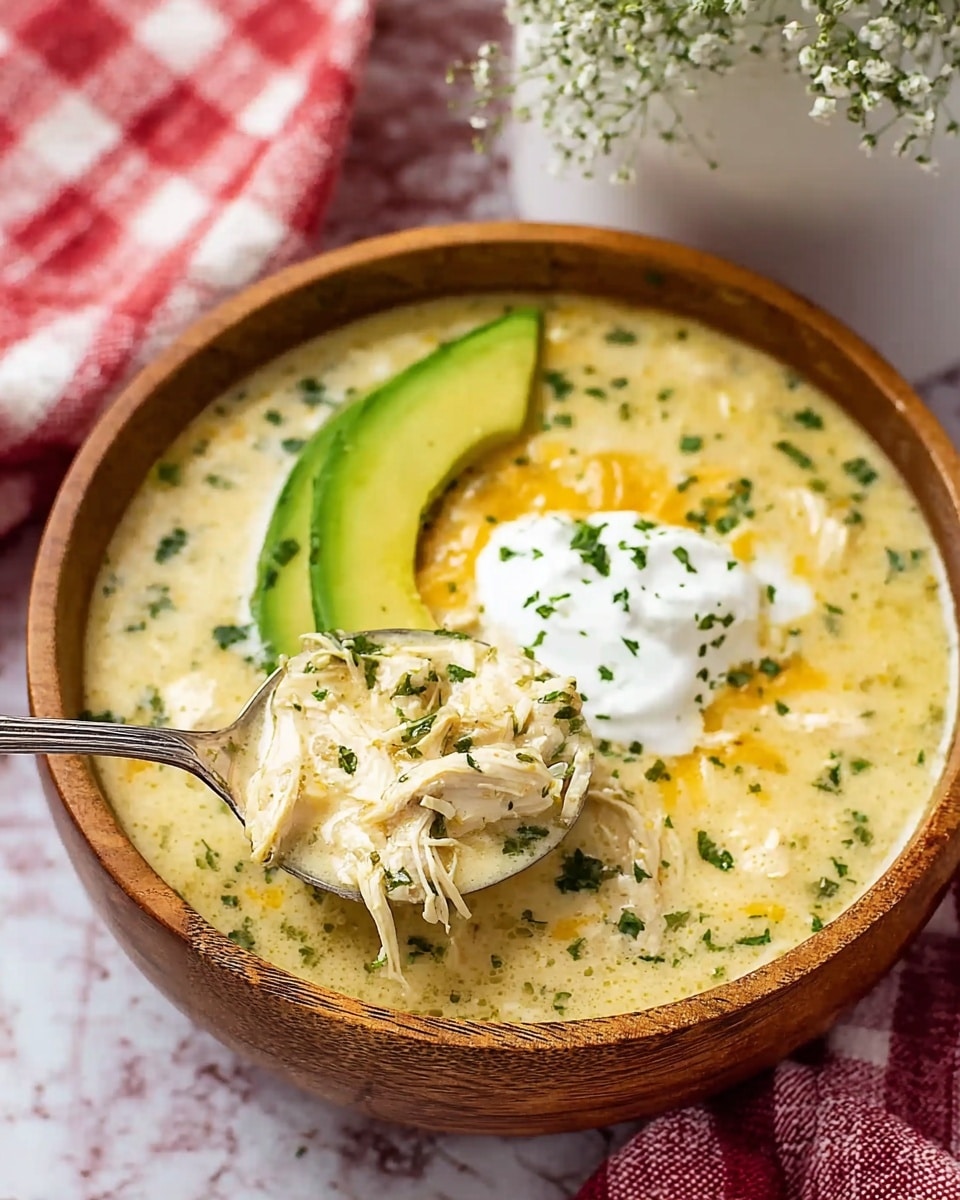 A wooden bowl filled with creamy yellow soup that has visible shredded white chicken pieces mixed in. On top of the soup, there are two dollops of white sour cream and sprinkled green chopped herbs. A thick slice of bright green avocado rests on the side inside the bowl. A silver spoon lifts from the bowl a portion of the soup showing chicken, sour cream, and herbs. The bowl sits on a white marbled surface with a red and white checkered cloth nearby. In the background, there is a blurred white vase with delicate white flowers. Photo taken with an iphone --ar 4:5 --v 7