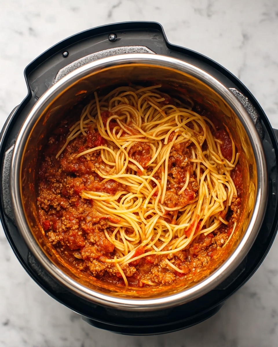 Inside a black electric pot with a shiny stainless steel inner layer, there is a mix of cooked spaghetti and thick red tomato meat sauce. The base layer is chunky ground meat in a rich, slightly oily tomato sauce with visible pieces of tomato. On top, light golden-yellow strands of spaghetti are spread unevenly, some tangled and some stretched out, mixing partly with the sauce below. The metal pot sits on a white marbled surface. photo taken with an iphone --ar 4:5 --v 7