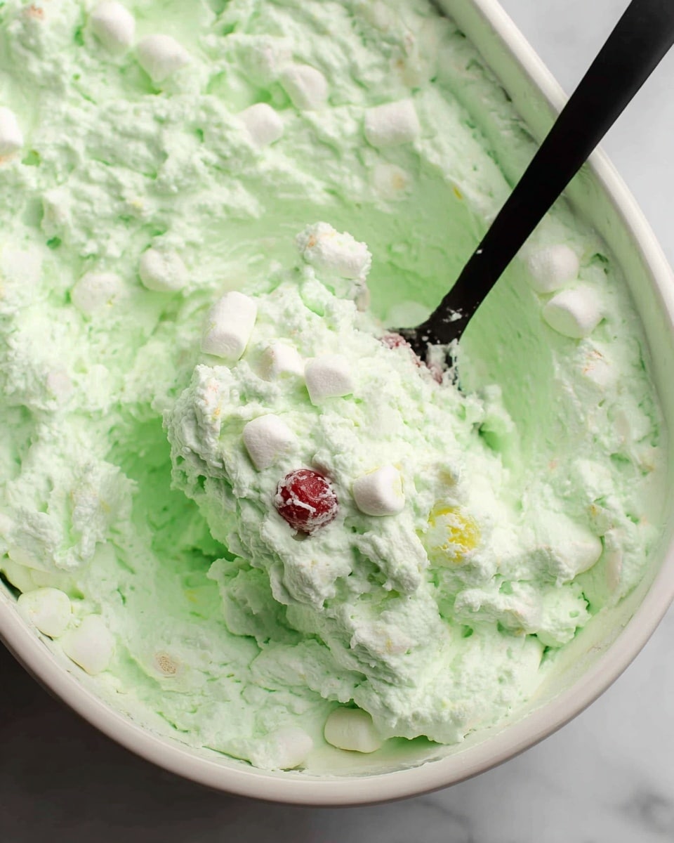 A close-up view of a white bowl filled with a light green, fluffy, and creamy mixture that contains small white marshmallows scattered throughout. The texture looks soft and slightly chunky with a mix of smooth and airy parts. The bowl is placed on a surface with a white marbled texture. In the background, there is a white tray that also contains the same green mixture, slightly out of focus, along with a white bowl filled with more white mini marshmallows. The colors are soft and pastel, giving a fresh and light appearance. photo taken with an iphone --ar 4:5 --v 7