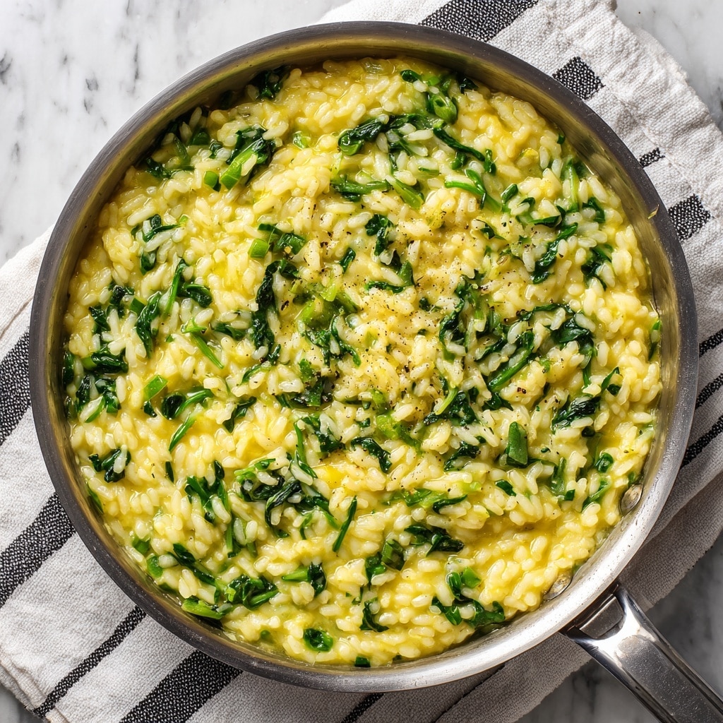 A close-up top view of a shiny metal pan filled with creamy yellow risotto mixed with bright green leafy vegetables throughout. The risotto has a soft and thick texture, with each grain of rice visible and gently coated in the sauce. The green vegetables are cut into small chunks and scattered evenly, adding vibrant color contrast. The pan is placed on a white and black striped cloth that lays on a white marbled surface. Photo taken with an iphone --ar 4:5 --v 7