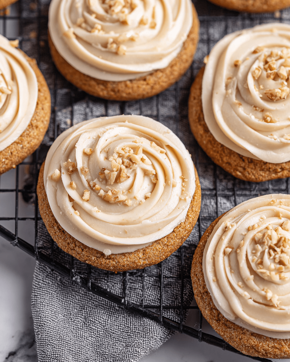 The image shows several cookies on a black cooling rack placed over a white marbled surface. Each cookie has two layers: the bottom layer is a golden-brown cookie with a slightly rough texture, and the top layer is creamy white frosting piped smoothly in a rose shape. Small pieces of chopped nuts are sprinkled lightly on the frosting and around the cookies on the cooling rack. The cookies are arranged closely together in a grid-like pattern. photo taken with an iphone --ar 4:5 --v 7