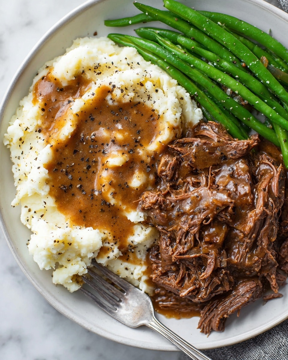 The image shows a white plate with three main parts: a layer of creamy white mashed potatoes topped with brown gravy sprinkled with black pepper on the left; on the right, a pile of shredded brown pot roast with a glossy texture and small bits of onion, a silver fork resting on it; above the roast, a neat row of bright green steamed green beans sprinkled lightly with black pepper; a small white bowl filled with a pale yellow side, possibly horseradish, is partially visible at the bottom left corner. The dish sits on a white marbled surface. Photo taken with an iphone --ar 4:5 --v 7