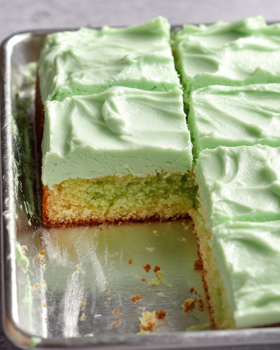 The image shows a rectangular green cake sliced into squares with one piece removed, revealing a two-layer look: a light green moist cake at the bottom and a thick, smooth, pale green frosting layer on top. The cake rests on a shiny metal tray with some cake crumbs scattered around. The frosting has a soft and creamy texture with subtle swirls on the top surface. The cut edges of the cake are clean and show the dense crumb of the cake inside. photo taken with an iphone --ar 4:5 --v 7