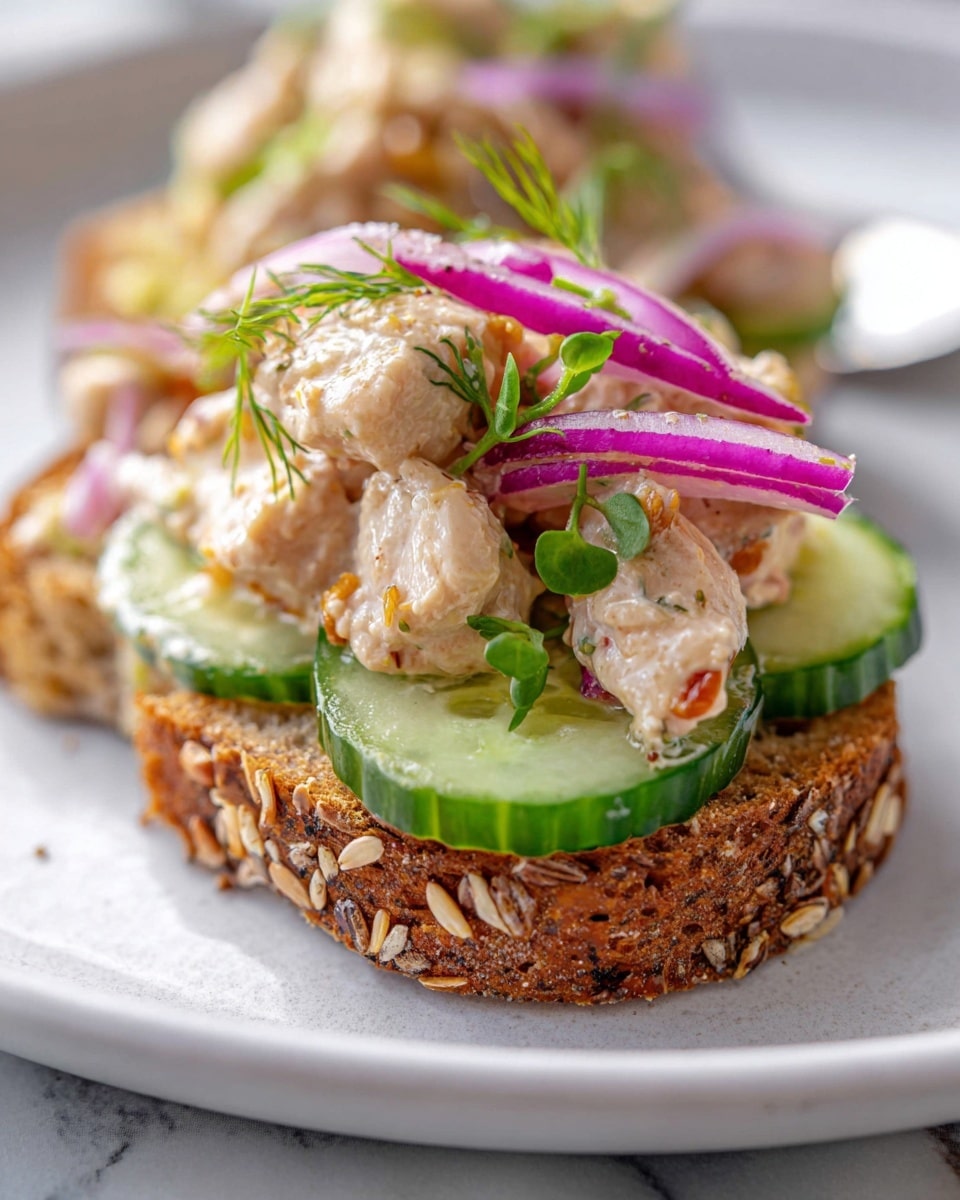 The dish shows an open-faced sandwich on a thick slice of toasted multigrain bread with visible oats on the crust as the bottom layer. On top of the bread rests fresh green leafy spinach, followed by a slice of red tomato. The next layer is chunky, seasoned shredded chicken, topped with thin slices of bright pink pickled red onions and a round green cucumber slice. A small sprig of fresh dill adds a touch of green garnish on top. The sandwich is placed on a white marbled surface, with blurred similar sandwiches in the background. Photo taken with an iphone --ar 4:5 --v 7