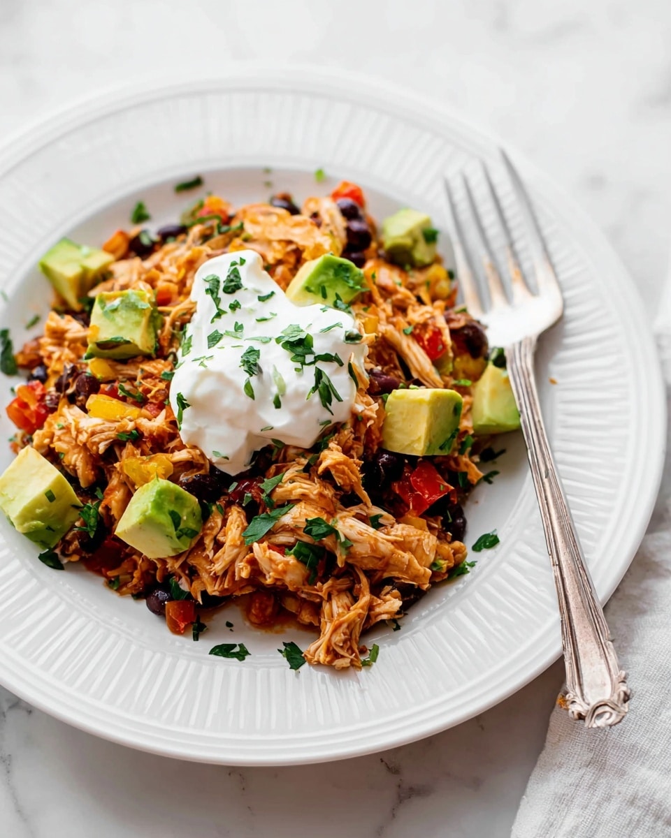 A white plate with ridged edges holds a colorful mixture as the main dish, featuring shredded orange-brown chicken combined with black beans and small pieces of red and yellow peppers. Around and within the mixture, there are bright green cubes of avocado and chopped green herbs sprinkled on top. At the center of the dish, a dollop of smooth white sour cream rests. To the side of the plate, a silver fork is placed partly on the food, on a white marbled surface. Photo taken with an iphone --ar 4:5 --v 7