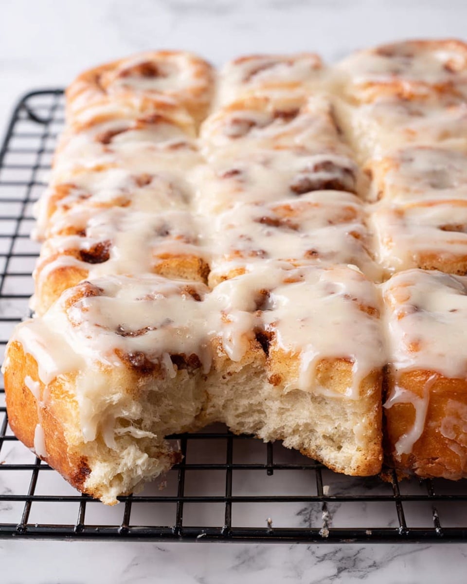 The image shows a square bun batch made of 12 pieces on a black cooling rack. The buns have a soft, airy texture with a golden-brown cinnamon swirl visible inside. Each bun is thickly covered with a creamy white glaze that looks smooth and slightly shiny, dripping onto the cooling rack at some points. The buns are closely placed together, showing the edges touching, with one piece pulled out a bit, showing the fluffy inside. The background is a white marbled surface. Photo taken with an iphone --ar 4:5 --v 7